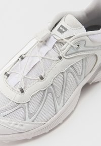Bílá sportovní obuv s síťovinovým povrchem, texturovaným designem, reflexními šedými prvky a pevně přiléhajícím elastickým zavazováním pro nastavitelný střih.