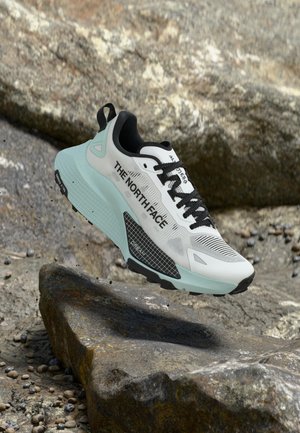 Fehér és világoskék The North Face sportcipő fekete fűzőkkel, sziklás terep felett lebegve.