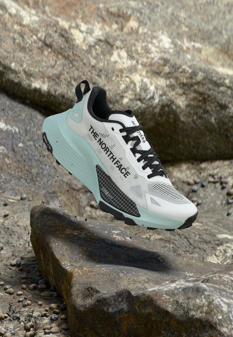 Λευκό και ανοιχτό μπλε αθλητικό παπούτσι The North Face με μαύρα κορδόνια που αιωρείται πάνω από πετρώδη έδαφος.