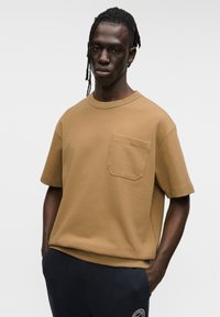 Καφέ κοντομάνικη μπλούζα με στρογγυλή λαιμόκοψη και τσέπη. Λεία υφή, άνετη εφαρμογή, σε συνδυασμό με σκούρο παντελόνι. Απλός σχεδιασμός.