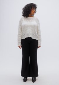 Λευκό, πλεκτό τοπ με μακρύ μανίκι και σχέδιο, συνδυασμένο με μαύρο παντελόνι φαρδύ με πέτα και μποτάκια στον αστράγαλο. Λευκό, ουδέτερο φόντο.