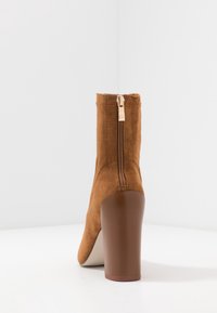 Bottine en suede marron avec une fermeture éclair à l'arrière et un talon épais et haut sur un fond minimaliste blanc.