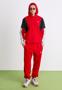 Persoană îmbrăcată cu un hanorac Nike roșu, negru și gri, cu pantalonii de trening roșii asortați și adidași albi, stând pe un podea verde, pe un fundal de perete alb.