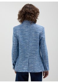 Personne aux cheveux bouclés vue de dos portant un blazer bleu en tweed à manches longues et un pantalon sombre sur un fond uni.