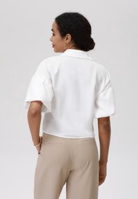 White, short-sleeved blouse with a relaxed fit and textured fabric; paired with beige tailored trousers featuring back pockets.
