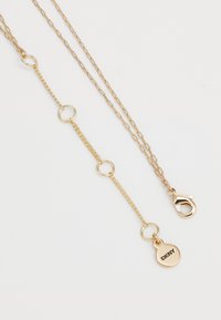 Collier en chaîne plaqué or avec de petits maillons ronds, un fermoir mousqueton et une étiquette gravée "DKNY". Texture lisse, design élégant.