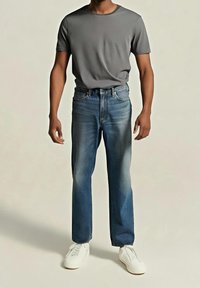 Homme portant un t-shirt gris uni à manches courtes, un jean bleu délavé droit et des baskets blanches, debout devant un fond neutre.