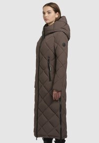 Brun lang pufferjakke med diamantmønstret quiltning, høj krave og sidelukninger. Har et brandet patch på ærmet og hætten.