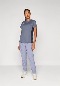 Jauna sieviete stāv, tērpusies pelēkā īsām piedurknēm sporta krekla, gaiši purpura jogger biksēs un baltās sporta apavos pret vienkāršu fona.