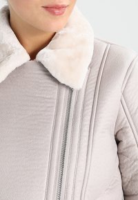 Chaqueta de cuero sintético de color rosa claro con un suave cuello de piel sintética color crema, que presenta detalles de costura vertical y un zipper plateado.