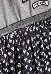 Haut gris avec des écussons portant les mots "YAY" et "AWESOME" au-dessus d'une jupe noire à motif floral. Présente une texture froncée et une ceinture argentée.