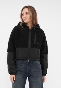 Giacca nera cropped che presenta un mix di pile testurizzato e tessuto liscio, con chiusura a zip, cappuccio e polsini elasticizzati.