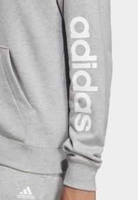 Šedá mikina s klokaní kapsou, žebrovanými manžetami a velkým bílým logem "adidas" na rukávu s dalšími brandingovými prvky na lemu.