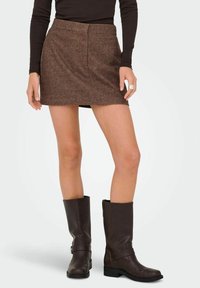 Vrouw in een bruine gestructureerde minirok, zwart langemouwtop, donkerbruine leren laarzen tot net onder de knie en een grote zilveren ring om haar rechterhand.