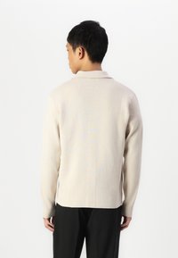Beige knit sweater with a collar, long sleeves, and a smooth texture. The back features a subtle pattern and a straight hemline.