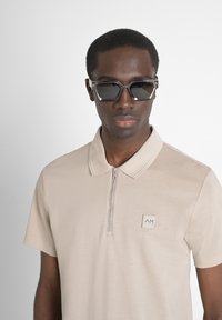 Polo beige de manga corta y cuello, con una cremallera frontal y un pequeño parche con logo. Gafas de sol negras con un marco geométrico.