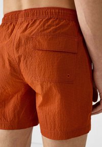 Shorts da bagno arancioni realizzati in tessuto leggero con motivo a rete, dotati di vita elastica e una tasca posteriore con chiusura a bottone.