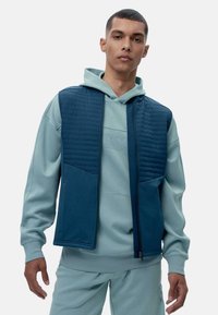 Tamsiai mėlyna siuvinėta liemenė su kontrastine tamsiai mėlyna apatine dalimi ir kišenėmis, uždėta ant šviesiai mėlynos dėvimos džemperio, demonstruojanti lygų, tekstūruotą paviršių.