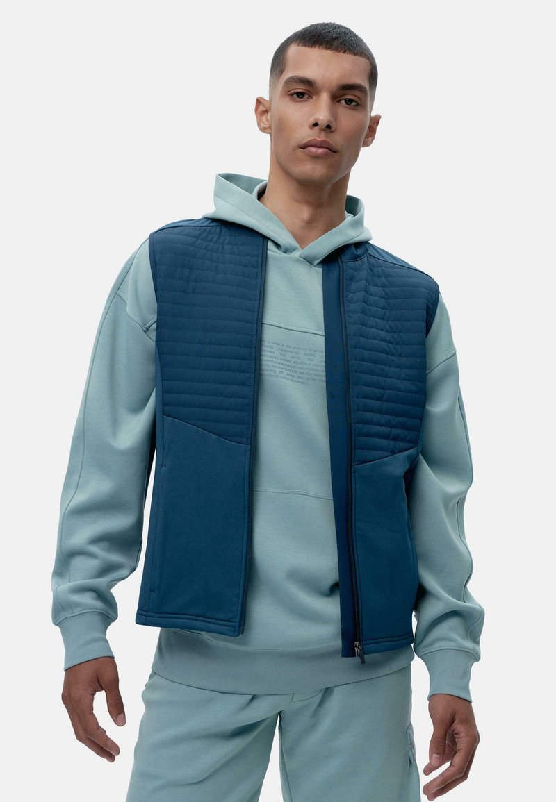 Tamsiai mėlyna siuvinėta liemenė su kontrastine tamsiai mėlyna apatine dalimi ir kišenėmis, uždėta ant šviesiai mėlynos dėvimos džemperio, demonstruojanti lygų, tekstūruotą paviršių.