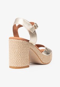 Tacones metálicos dorados con una plataforma trenzada beige. Cuenta con una correa ajustable en el tobillo y material texturizado. Diseño de punta cerrada.