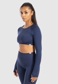 Młoda kobieta z długimi włosami w granatowej, długorękawowej bluzce crop top oraz legginsach z wysokim stanem, stojąca na jasnym tle.