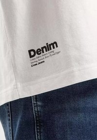 Biała bawełniana koszulka z czarną metką "Denim", zawierającą szczegóły dotyczące materiału i konstrukcji, w połączeniu z ciemnoniebieskimi dżinsami.