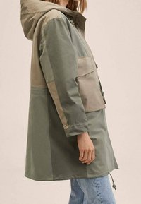 Veste légère à capuche en vert et beige, avec une poche avant et un ourlet ajustable. Confectionnée en tissu lisse avec une finition légèrement brillante.