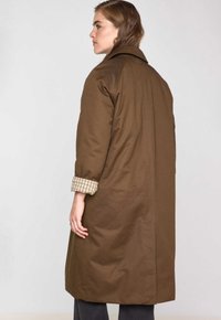 Donna dai capelli castano medi che indossa un lungo cappotto marrone con le maniche arrotolate che mostrano una fodera a quadri, rivolta a sinistra su uno sfondo semplice.