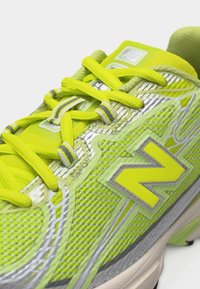 Φωτεινό κίτρινο-πράσινο αθλητικό παπούτσι με υφασμάτινο δίχτυ, αντανακλαστικές ασημένιες λεπτομέρειες, κορδόνια με ριγέ σχέδιο και μεγάλο λογότυπο "N" στο πλάι.