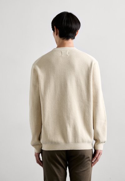 Pull en tricot de couleur crème clair avec un motif texturé, col rond et poignets côtelés, associé à un pantalon vert olive foncé.