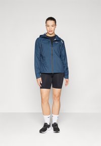 Marine waterdichte jas met capuchon, rits aan de voorkant en twee zijzakken, gecombineerd met zwarte shorts en zwarte sportschoenen.