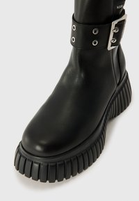 Black leather ankle boot featuring a chunky, ridged sole, adjustable buckle strap with silver hardware, and smooth texture.