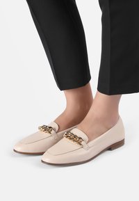 Beige Lederloafers mit einer goldenen Kettenakzent, flacher Sohle, mandelförmiger Zehenpartie und glatter Textur, getragen mit schwarzen, gekürzten Hosen.