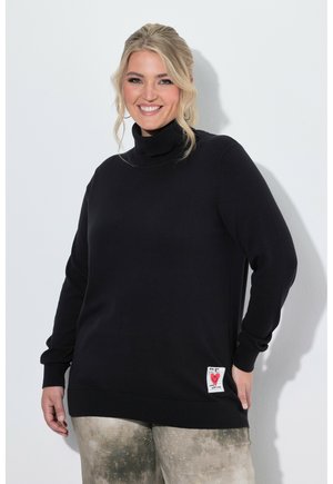 Smilende kvinde med blondt hår iført sort rullekravesweater og lyse mønstrede bukser står foran en hvid baggrund.