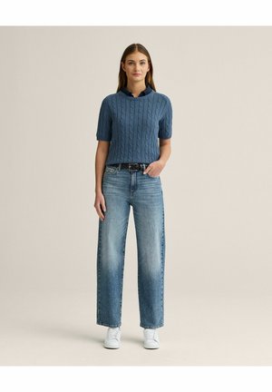 Kvinde iført blå kabelstrikket kortærmet sweater, blå skjorte med krave, brede jeans og hvide sneakers, stående mod en lys baggrund.