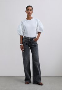 Top blanco con mangas abullonadas combinado con jeans gris de pierna ancha y alta cintura. El material parece suave con una textura lisa. Sandalias visibles.