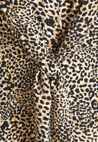 Skjorta i leopardmönster med en knuten design vid midjan, gjord av lättviktsmaterial med svarta och gyllene fläckar mot en krämfärgad bakgrund.