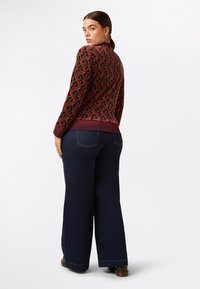 Giacca bomber bordeaux con motivo, dotata di colletto e polsini a coste, abbinata a jeans a gamba larga blu scuro e scarpe marroni. Dettagli di cuciture sottili visibili.