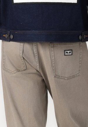 Pantalones beige con dos bolsillos traseros, uno de ellos con una pequeña placa en blanco y negro, usados con una chaqueta azul oscuro visible en la cintura.