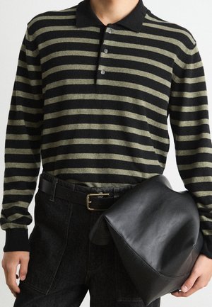 Persoon draagt een zwart-olijfgroen gestreept poloshirt met lange mouwen en een zwarte broek, houdt een zwartleren etui in één hand vast.