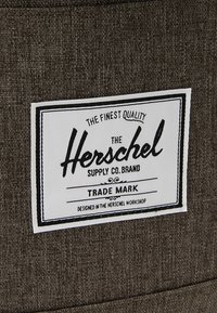 Étiquette rectangulaire blanche de la marque Herschel Supply Co. avec texte noir cousue sur un tissu marron foncé texturé.