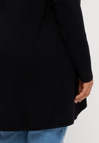 Black long-sleeve top with a rounded hem, made of soft fabric. Sleeves have a subtle cuff; worn with light blue jeans.