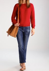 Maglione rosso a maglia texturizzata con scollo rotondo, abbinato a jeans blu a gamba dritta e sandali marroni. Tiene in mano una borsa in suede color tortora.