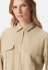 Beige overhemdd met een kraag, klepzakken en een gladde textuur. Heeft een relaxte pasvorm en goudkleurige knopen.