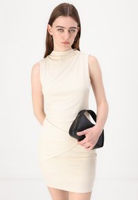 Cream-colored ribbed mini dress with a high neckline and sleeveless design, featuring a twisted waist detail. Model holds a small black bag.