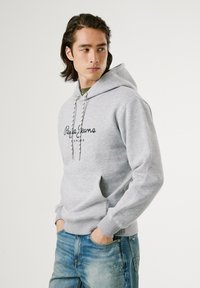 Sudadera gris claro con cordón ajustable, estampado del logo en la parte frontal y bolsillo de canguro. Combinada con jeans azules desgastados. Textura de tela suave.