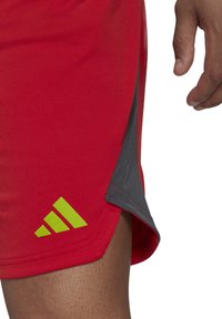 Rote Sportshorts mit einem grauen Seitenpanel, das ein leuchtend grünes Adidas-Logo auf der unteren linken Seite zeigt. Der Stoff wirkt glatt und leicht.