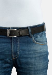 Crni kožni remen sa srebrnom metalnom kopčom, nošen preko plavih traperica. Traperice imaju klasičan dizajn s pet džepova i narančastim šavovima.