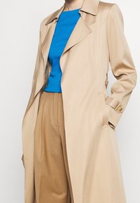 Trench beige con cuello notch, bolsillos laterales y botones de acento dorado, superpuesto sobre una blusa azul y pantalones marrones. Textura suave.