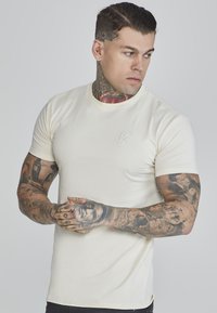 Tricou crem cu mânecă scurtă, cu guler rotund texturat, prezentând un logo brodat discret pe piept. Materialul pare neted, cu o ușoară elasticitate.